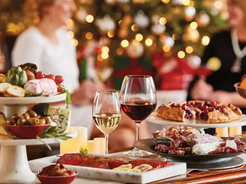 A photo of wine for Christmas with two glasses of wine sitting on a table surrounded by charcuterie and other appetizers with holiday decorations and people in the background