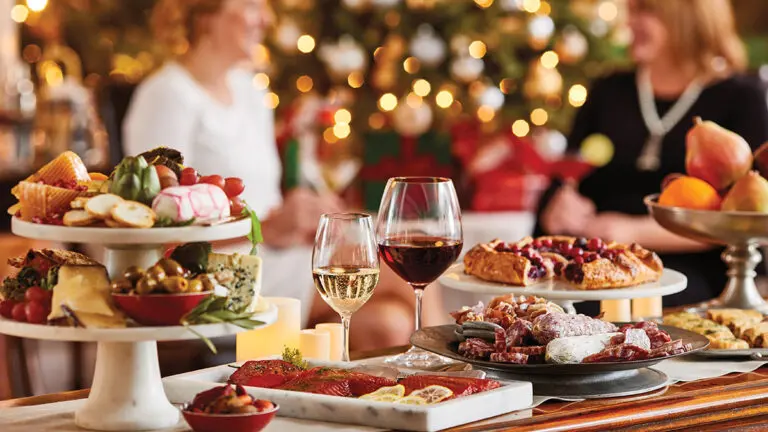 A photo of wine for Christmas with two glasses of wine sitting on a table surrounded by charcuterie and other appetizers with holiday decorations and people in the background