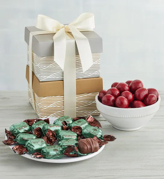 Gifts under $30 for Christmas with two gift boxes stacked on top of each other next to a bowl of chocolate covered cherries and a plate of chocolates wrapped in foil.