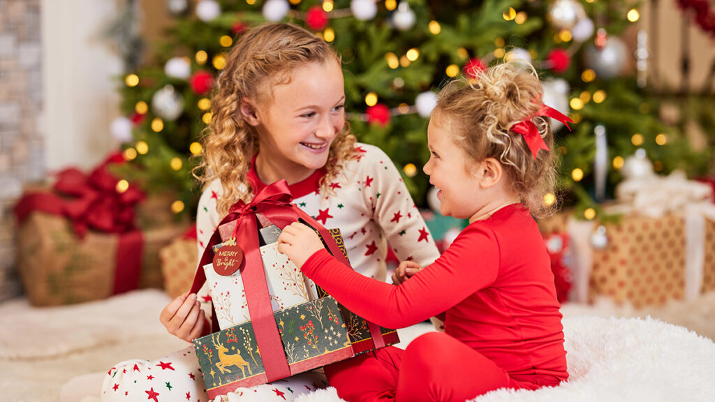 Christmas gifts under $30 with two girls smiling at each other as they open a Christmas gift under a tree.