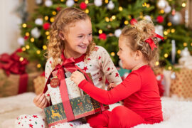 Christmas gifts under $30 with two girls smiling at each other as they open a Christmas gift under a tree.
