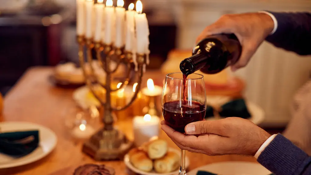 A photo of Hanukkah wine with a hand pouring wine into a glass with a menorah in the background