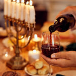 A photo of Hanukkah wine with a hand pouring wine into a glass with a menorah in the background