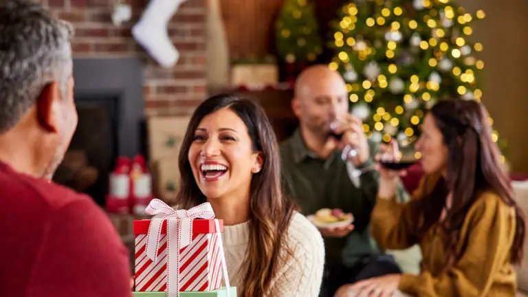 A photo of holiday shopping tips with a woman giving a man a wrapped gift with two people talking and drinking wine in the background
