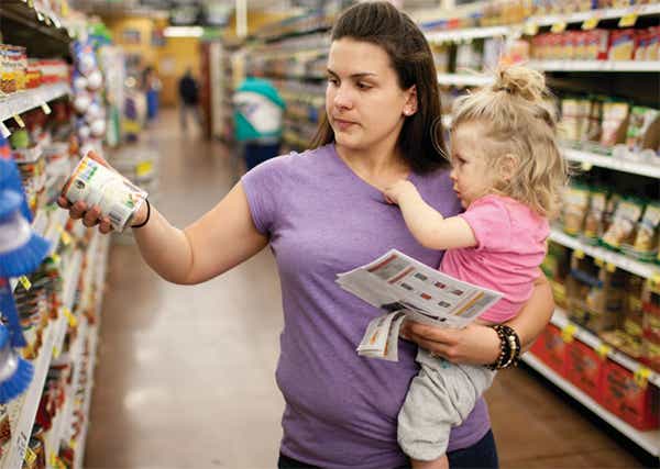A photo of food bank with a woman holding a child while they look at cans of food.