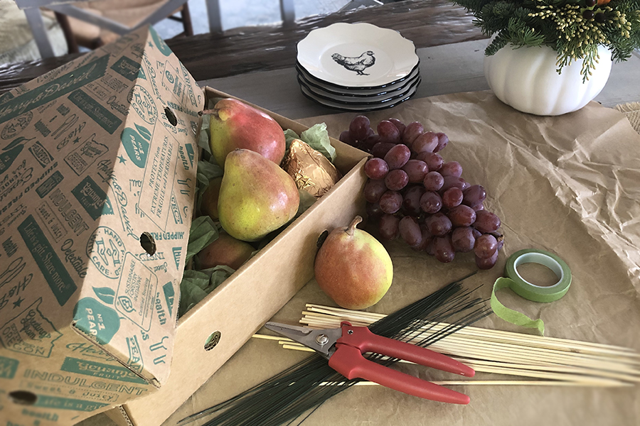 A photo of a pear tablescape with a box of pears and a bunch of grapes on a table with skewers and a pair of clippers