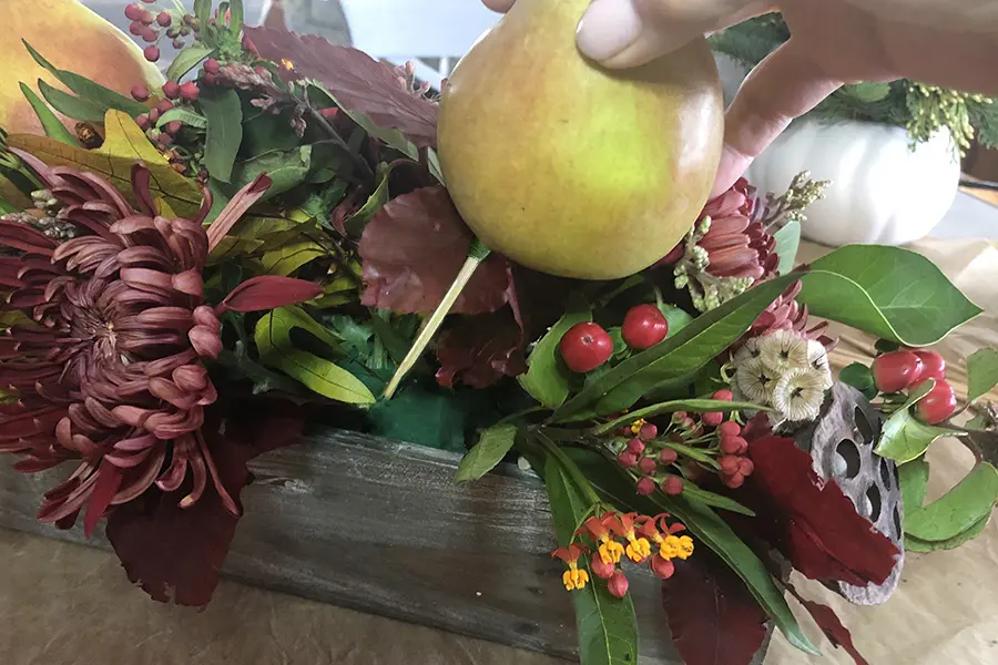 A photo of a pear tablescape with a pear on a stick being stuck into a box full of other greenery