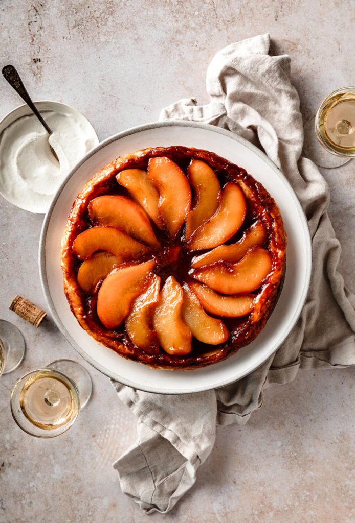 A photo of a pear tart on a plate surrounded by a bowl and two glasses of wine