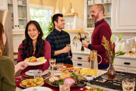 A photo of thanksgiving hosts with a group of people gathered in a kitchen sharing food and wine