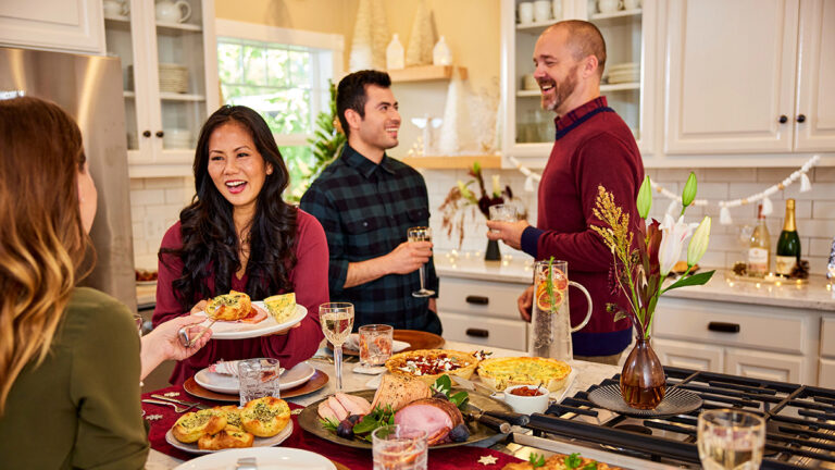 A photo of thanksgiving hosts with a group of people gathered in a kitchen sharing food and wine