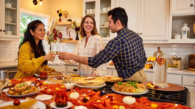 A photo of Thanksgiving tips with three people in a kitchen clinking wine glasses with a spread of food next to them