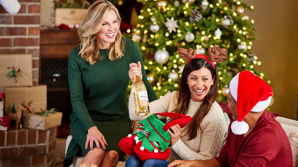 A photo of the ultimate wine gift basket with a woman opening up a Christmas present of wine with two other people sitting next to her and a Christmas tree in the background