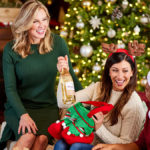 A photo of the ultimate wine gift basket with a woman opening up a Christmas present of wine with two other people sitting next to her and a Christmas tree in the background
