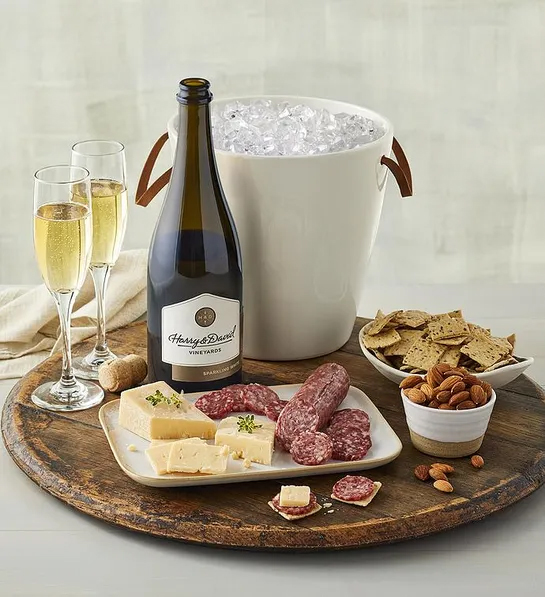 A bottle of wine next to a wine chiller full of ice and two glasses of sparkling wine on a tray surrounded by charcuterie and cheese