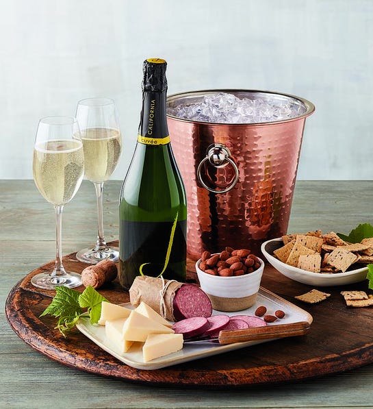 A photo of a wine basket with a platter holding a wine chiller full of ice, a bottle of sparkling wine, two wine glasses full of sparkling wine, and charcuterie surrounding it.