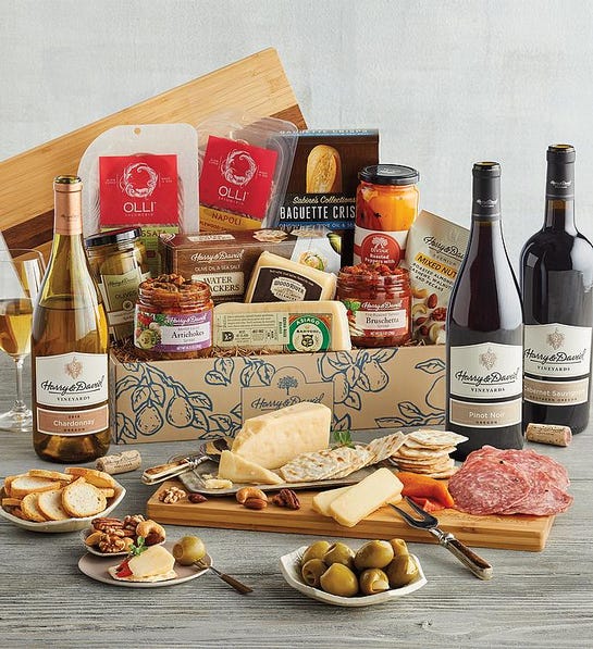 A photo of a wine basket full of charcuterie, cheese, and crackers surrounded by the same ingredients displayed on a cutting board and plates and three bottles of wine.