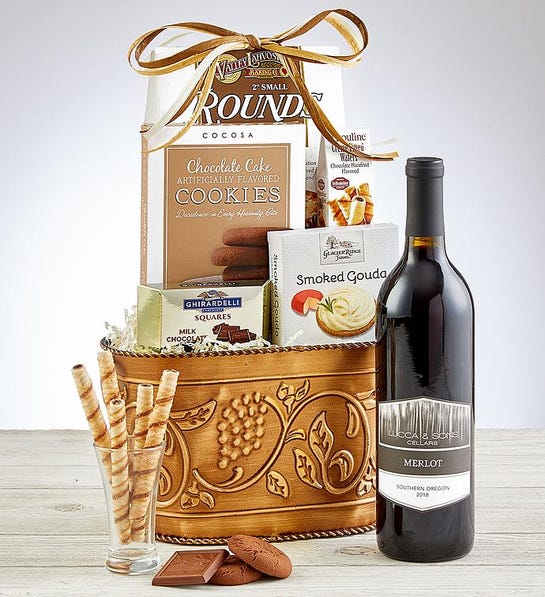 A photo of a wine basket made out of stamped tin full of crackers, cookies and chocolate with a bottle of wine in front and a few cookies next to it.