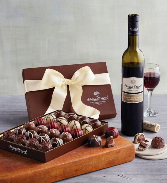 A photo of a wine basket with a cutting board holding a box of chocolates and a bottle of red wine in the background.