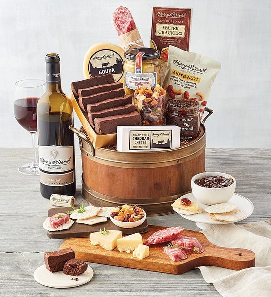 A photo of a wine basket shaped like a wine barrel full of slices of cake, cheese, crackers, and other snacks with a cutting board and several plates full of the same items in front and a bottle of wine in the background.