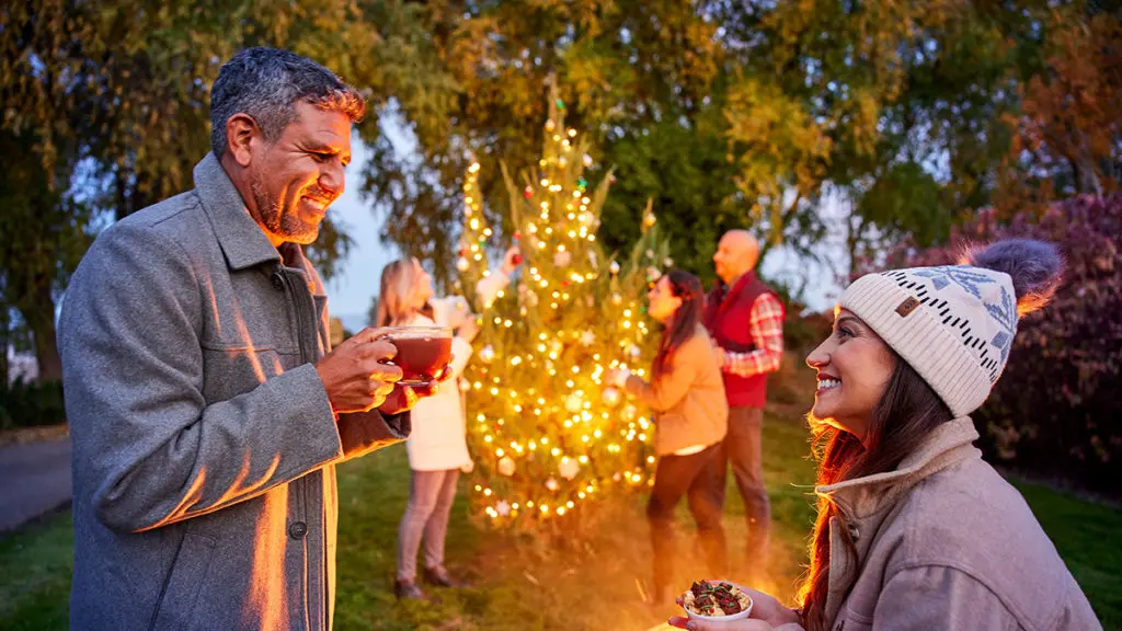 A photo of winter solstice with two people smiling at each other with cups of coco in hand and three people in the background decorating a Christmas tree outside