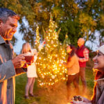 A photo of winter solstice with two people smiling at each other with cups of coco in hand and three people in the background decorating a Christmas tree outside