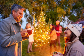 A photo of winter solstice with two people smiling at each other with cups of coco in hand and three people in the background decorating a Christmas tree outside