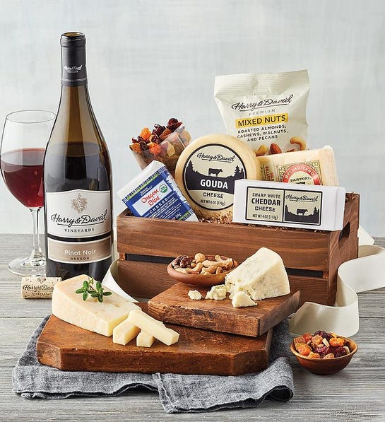 A photo of birthday gift ideas with a box full of cheese and snacks next to a bottle of wine and full glass with a cutting board holding more cheese and nuts in front.