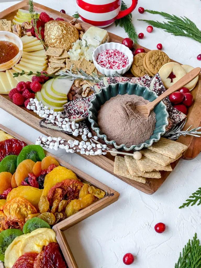 A photo of a Christmas charcuterie board with cheese, fruit, meat and nuts on a board surrounding a ramekin of hot cocoa powder