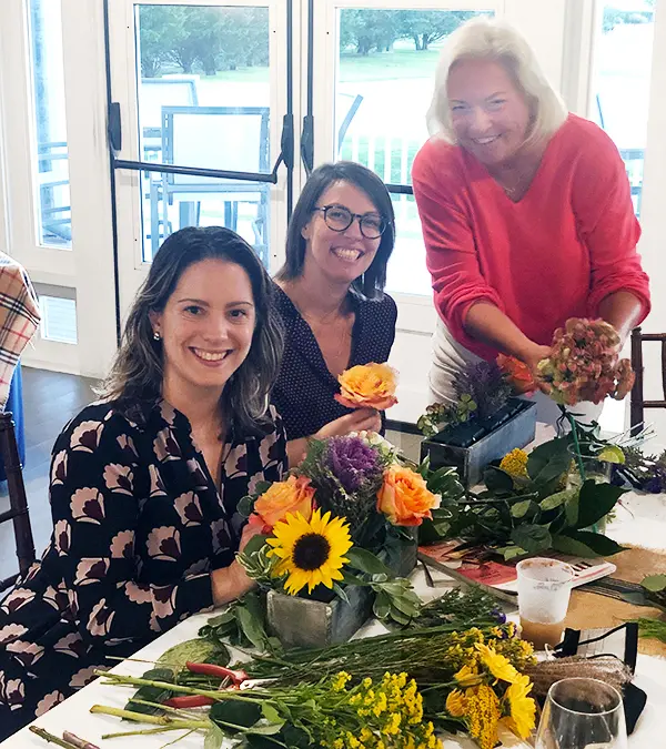 A photo of a craft cocktail with three women at a table arranging flowers.
