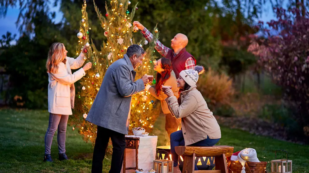 A photo of facts about Christmas with a group of people decorating a Christmas tree outside and drinking coco.