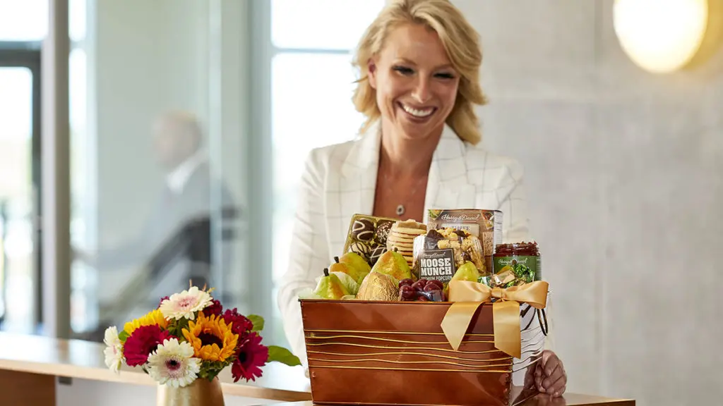 A photo of employee appreciation day with a woman at a desk smiling and looking at a gift basket full of fruit and other snacks.