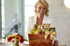 A photo of holiday gifts for employees with a woman at a desk smiling and looking at a gift basket full of fruit and other snacks.