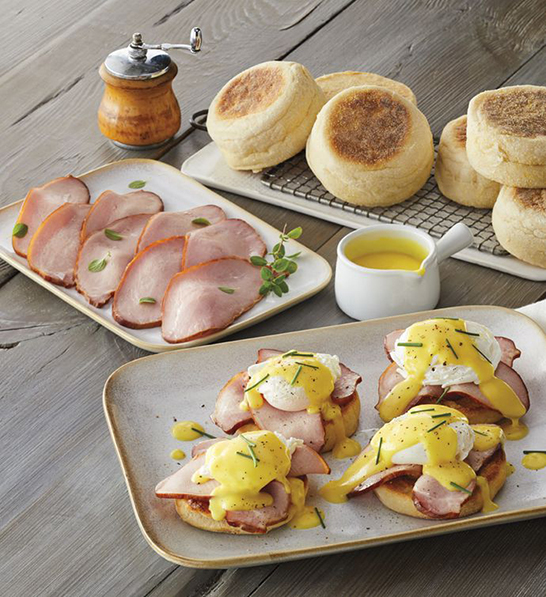 A photo of holiday gifts for employees with a platter of eggs Benedict and a platter of ham and a tray of English muffins in the background.