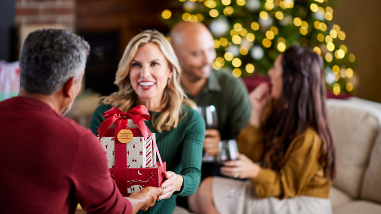 A photo of Christmas gift ideas with a woman handing someone a holiday gift with two people laughing in the background
