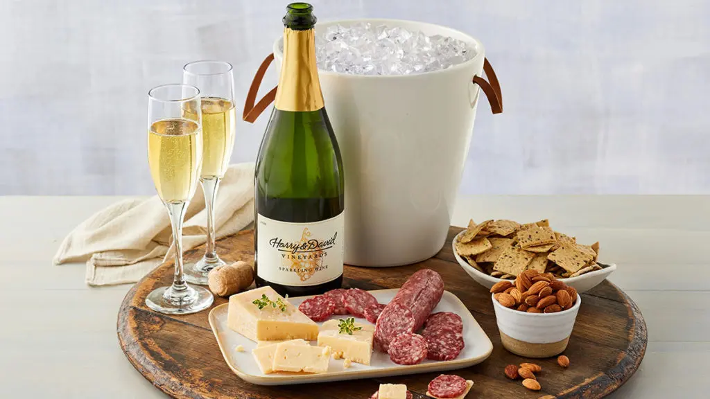 A photo of sparkling wine with an open bottle of wine next to a bucket full of ice and two flutes of wine all set behind a plate of meat and cheese with ramekins of nuts and crackers.