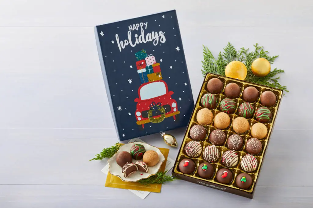 A photo of truffles with a box containing 24 truffles of different colors with a plate of the four of the same truffles next to it with the lid of the box propped up next to the box.
