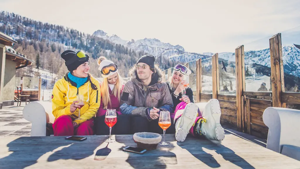 A photo of après ski with a group of people wearing snow gear sitting on a couch drinking wine outside with snow covered mountains in the background.