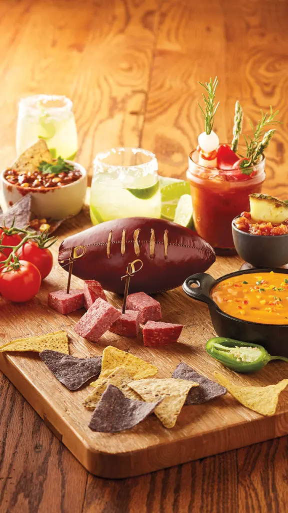 A photo of the big game with an array of finger foods arranged around a football shaped salami with several cocktails and other chips and dip.