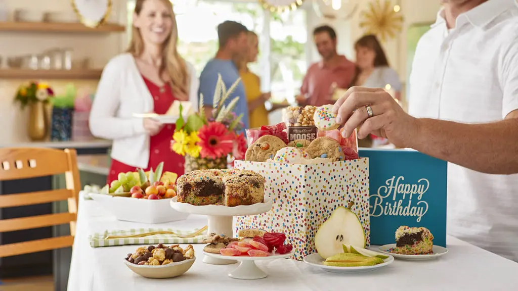 A photo of birthday party trends with a table full of birthday presents, cake, and cookies with people in the background