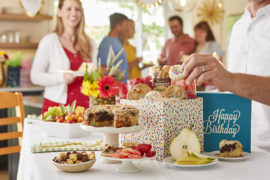 A photo of birthday party trends with a table full of birthday presents, cake, and cookies with people in the background