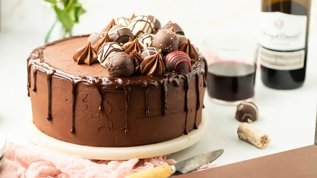 A photo of chocolate cake recipe with a chocolate cake on a cake stand covered in icing and truffles with a bottle of wine in the background