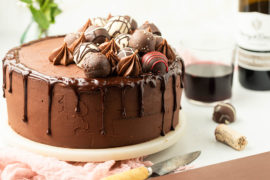 A photo of chocolate cake recipe with a chocolate cake on a cake stand covered in icing and truffles with a bottle of wine in the background