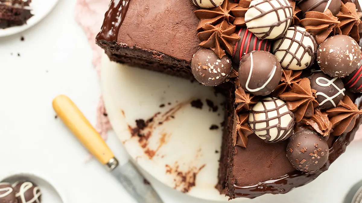 A photo of chocolate cake recipe with an above shot of a cake that's been sliced into that's covered in truffles and icing.