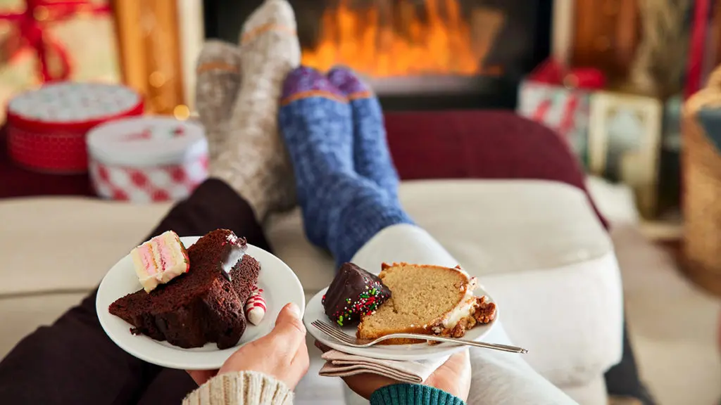 A photo of comfort foods with two pairs of feet in socks resting on a stool with a fire in the background and two hands holding two plates of cake in the foreground.