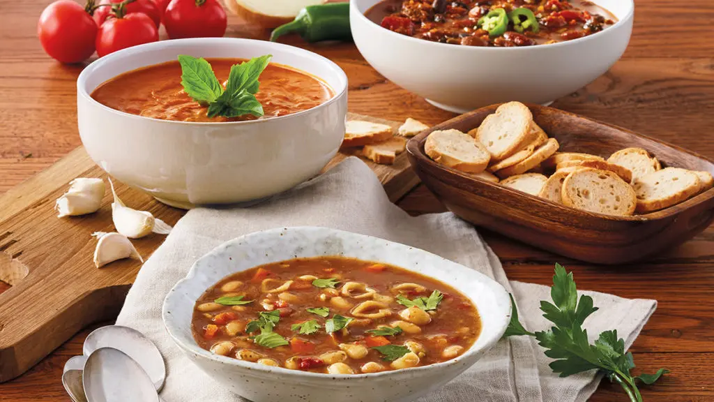 A photo of comfort foods with three bowls of different kinds of soup with a bowl of sliced bread.