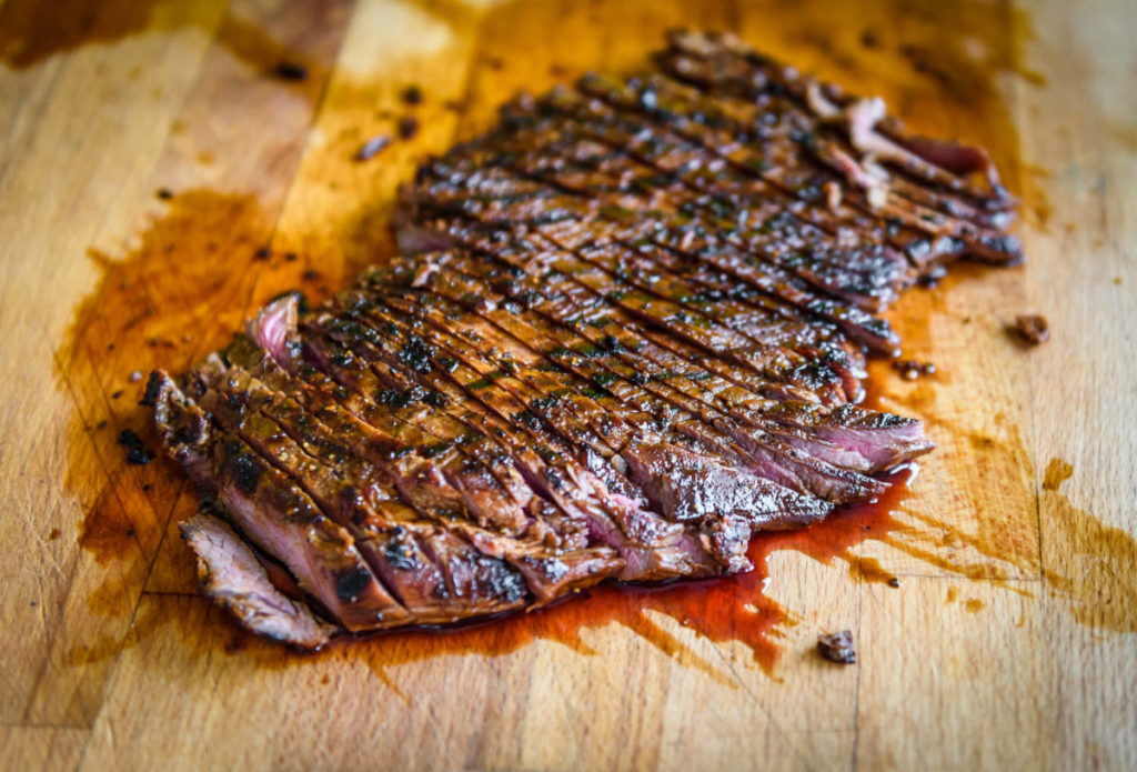 A photo of cuts of beef with a cutting board supporting a flank steak that has been cooked and sliced thinly.