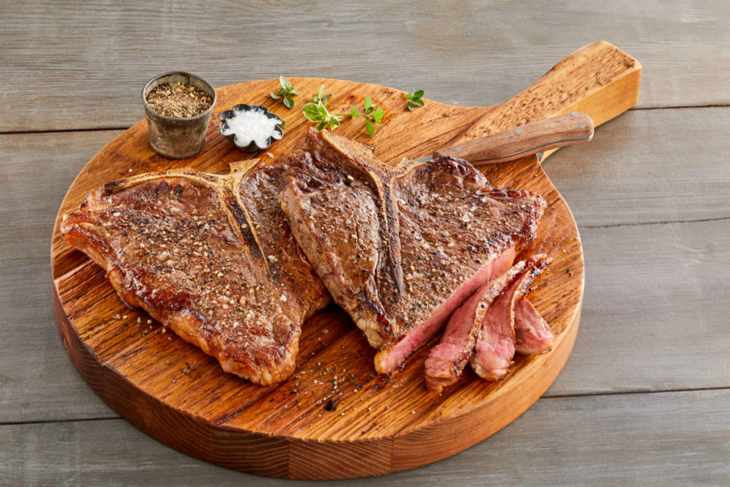 A photo of cuts of beef with a cutting board supporting two cooked t-bone steaks.