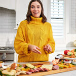 A photo of food trends with a woman standing in a kitchen with an array of charcuterie, cheese, and fruit on a table in front of her.