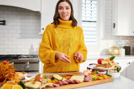 A photo of food trends with a woman standing in a kitchen with an array of charcuterie, cheese, and fruit on a table in front of her.