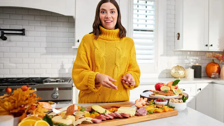 A photo of food trends with a woman standing in a kitchen with an array of charcuterie, cheese, and fruit on a table in front of her.